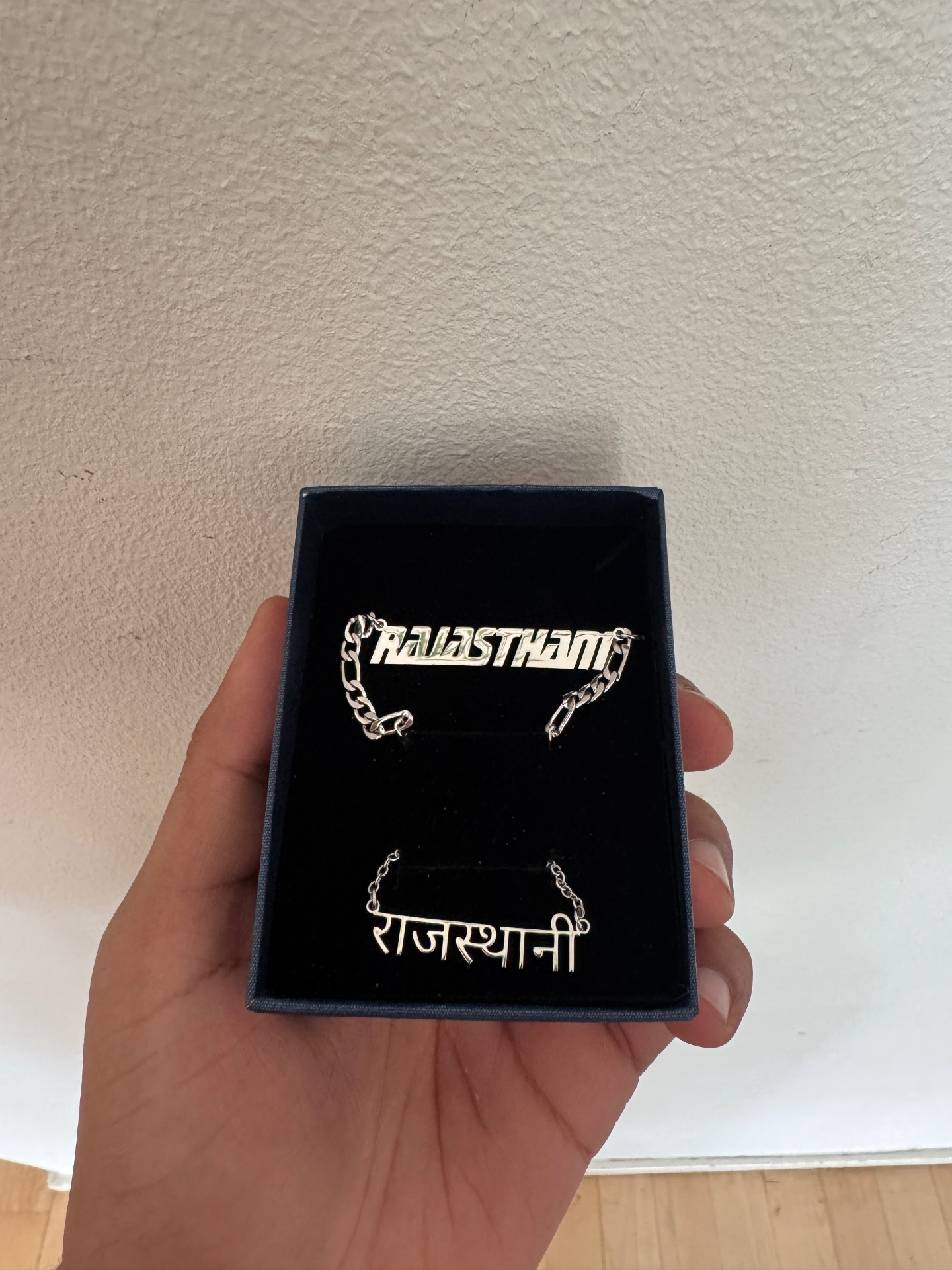 Two silver nameplate necklaces reading RAJASTHANI in Latin and Devanagari script in a jewellery box